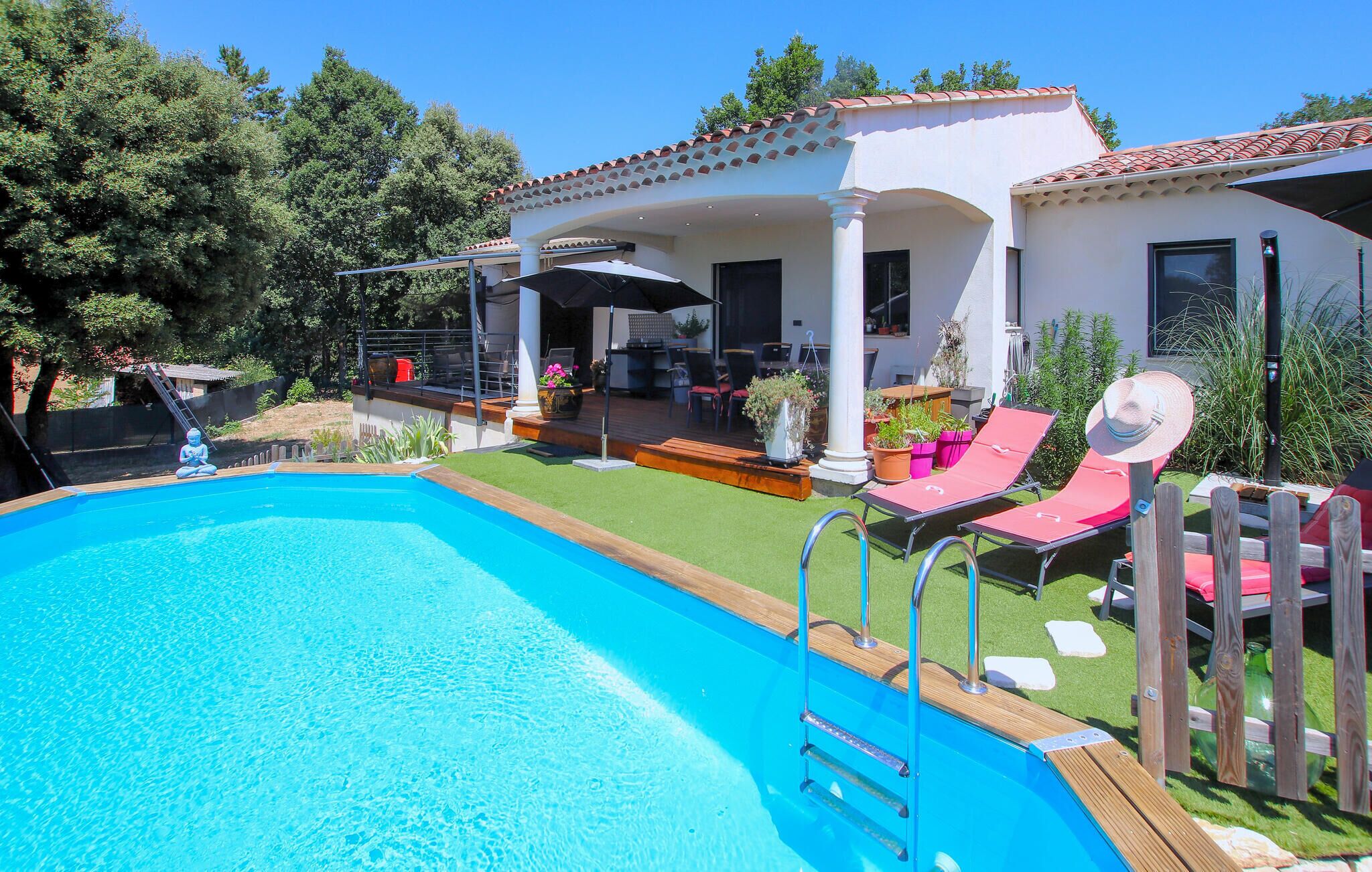 A swimming pool with a wooden deck, a covered patio area with outdoor furniture, and a white building with a red-tiled roof.