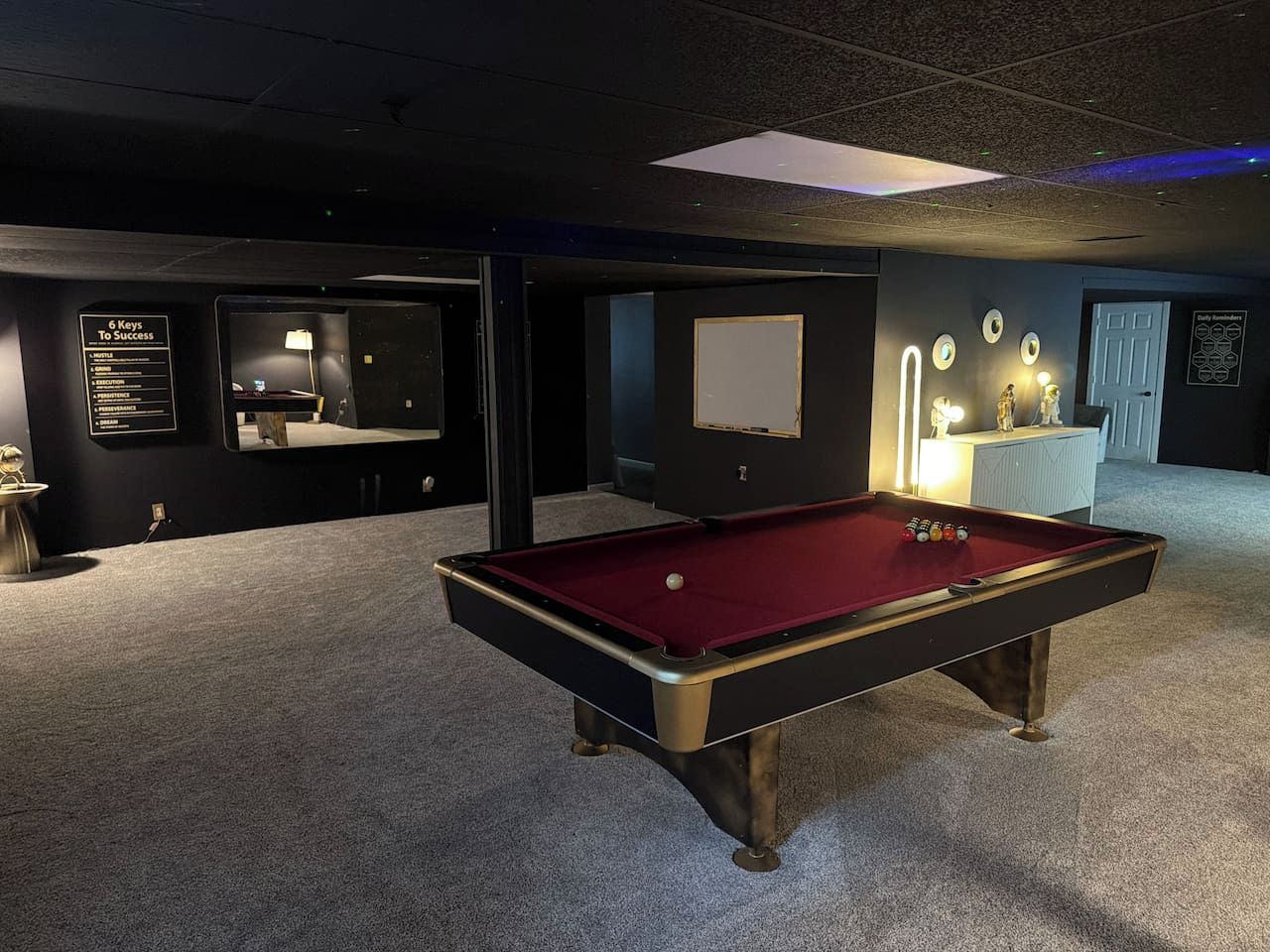 Une table de billard dans une pièce dont le sol est recouvert d’une moquette, avec une porte blanche et un mur orné d’une enseigne.