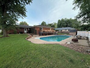 A backyard with a swimming pool, a brick house, and a fenced area with outdoor furniture.