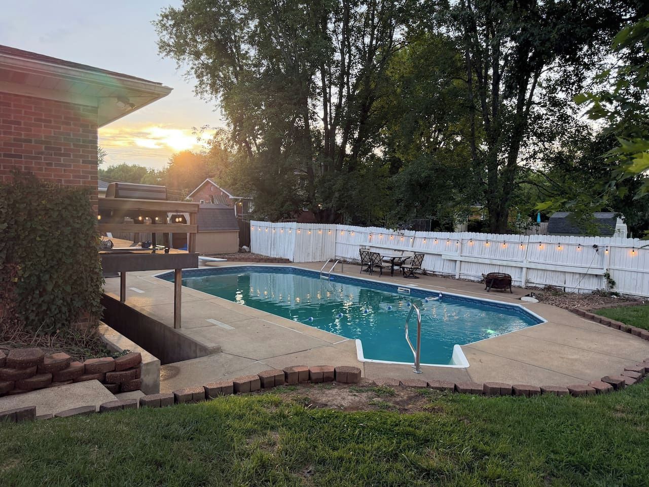 Un jardin avec une piscine, une terrasse dotée de mobilier d’extérieur et une clôture blanche.