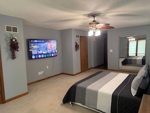 A bedroom with a bed, a ceiling fan, a TV mounted on the wall, and a window with a view.