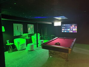 A pool table in a dimly lit room with green ambient lighting, a sofa, and a television displaying a sports channel.