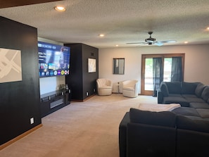 A living room with a flat-screen TV, two armchairs, a sofa, a ceiling fan, and a door leading outside.