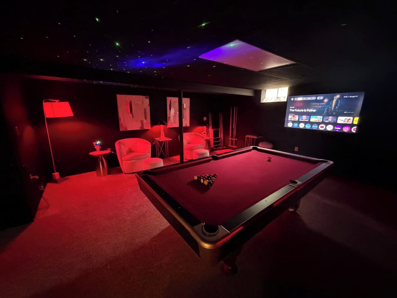 Une pièce faiblement éclairée, avec une table de billard, un éclairage d’ambiance rouge et un écran diffusant une chaîne de télévision.