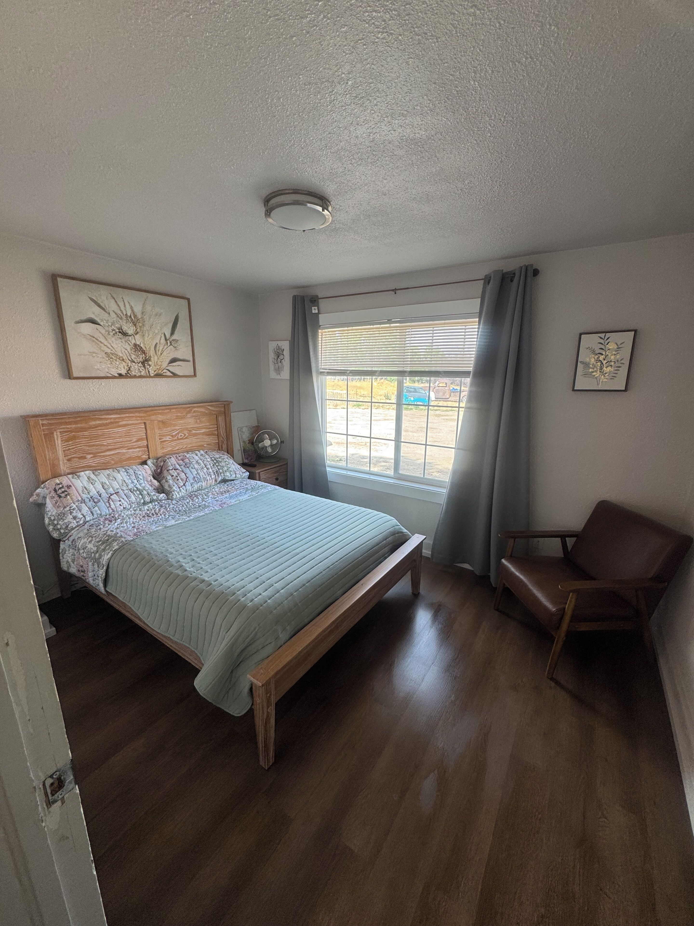 A bedroom with a bed, a chair, a window with curtains, and artwork on the walls.