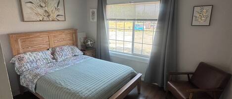 A bedroom with a bed, a chair, a window with curtains, and artwork on the walls.