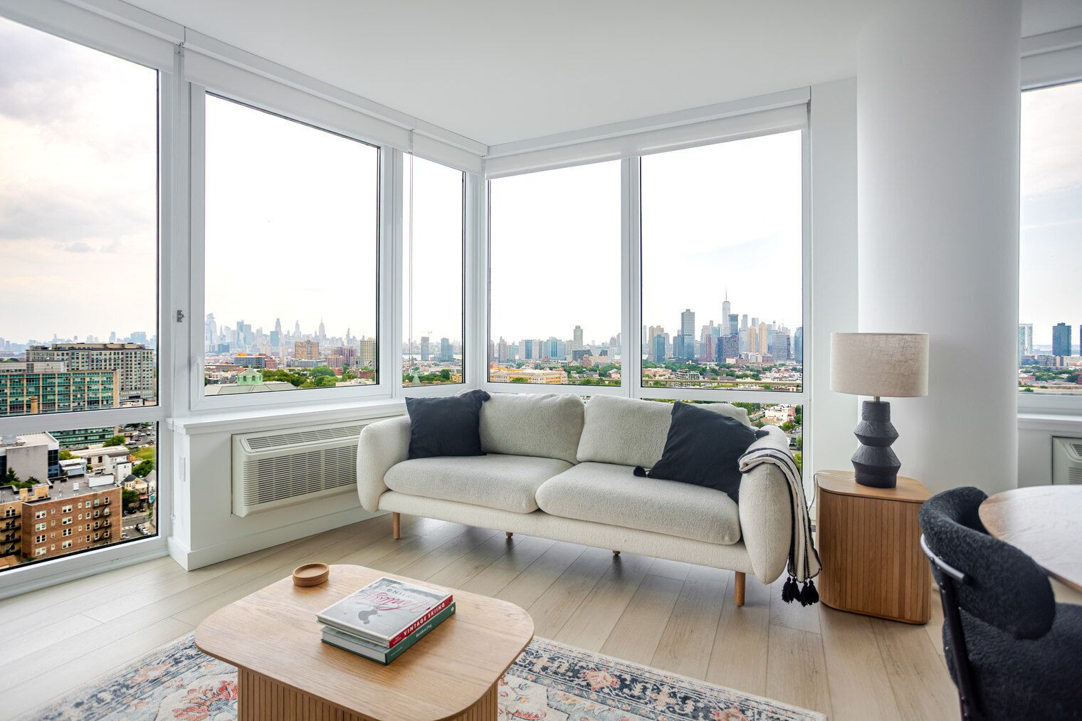 A modern living room with a sofa, a coffee table, and a lamp. Large windows offer a city view.