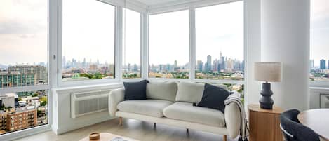 A modern living room with a sofa, a coffee table, and a lamp. Large windows offer a city view.