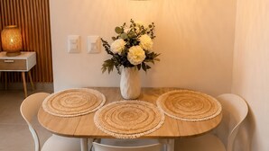 Spiseområde med et rundt bord, hvide stole, en vævet dug, en vase med blomster og en hængende lampe.