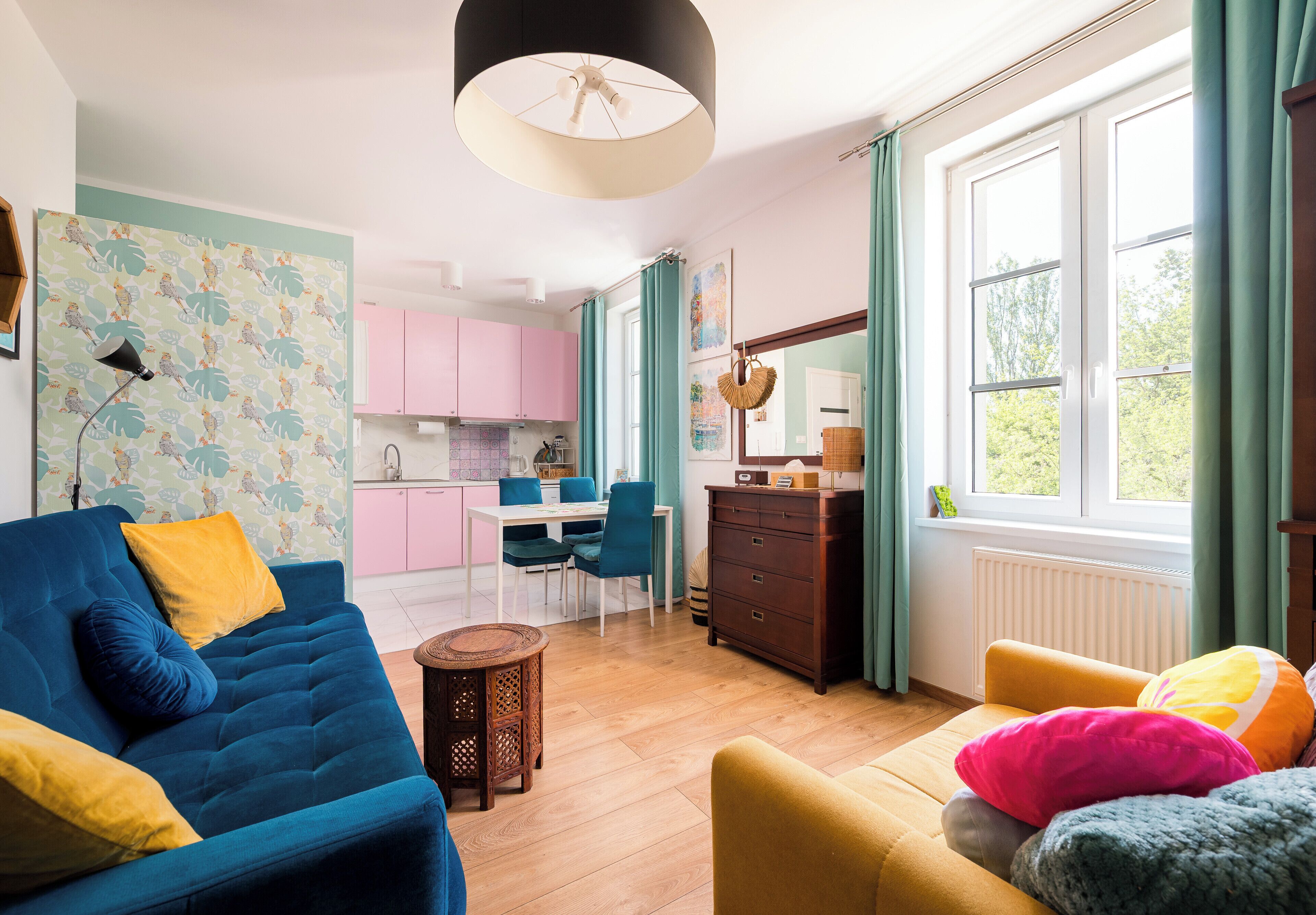 A modern living room with a blue sofa, yellow cushions, a yellow armchair, and a small round table. There is a kitchen area with pink cabinets and a dining table with chairs.