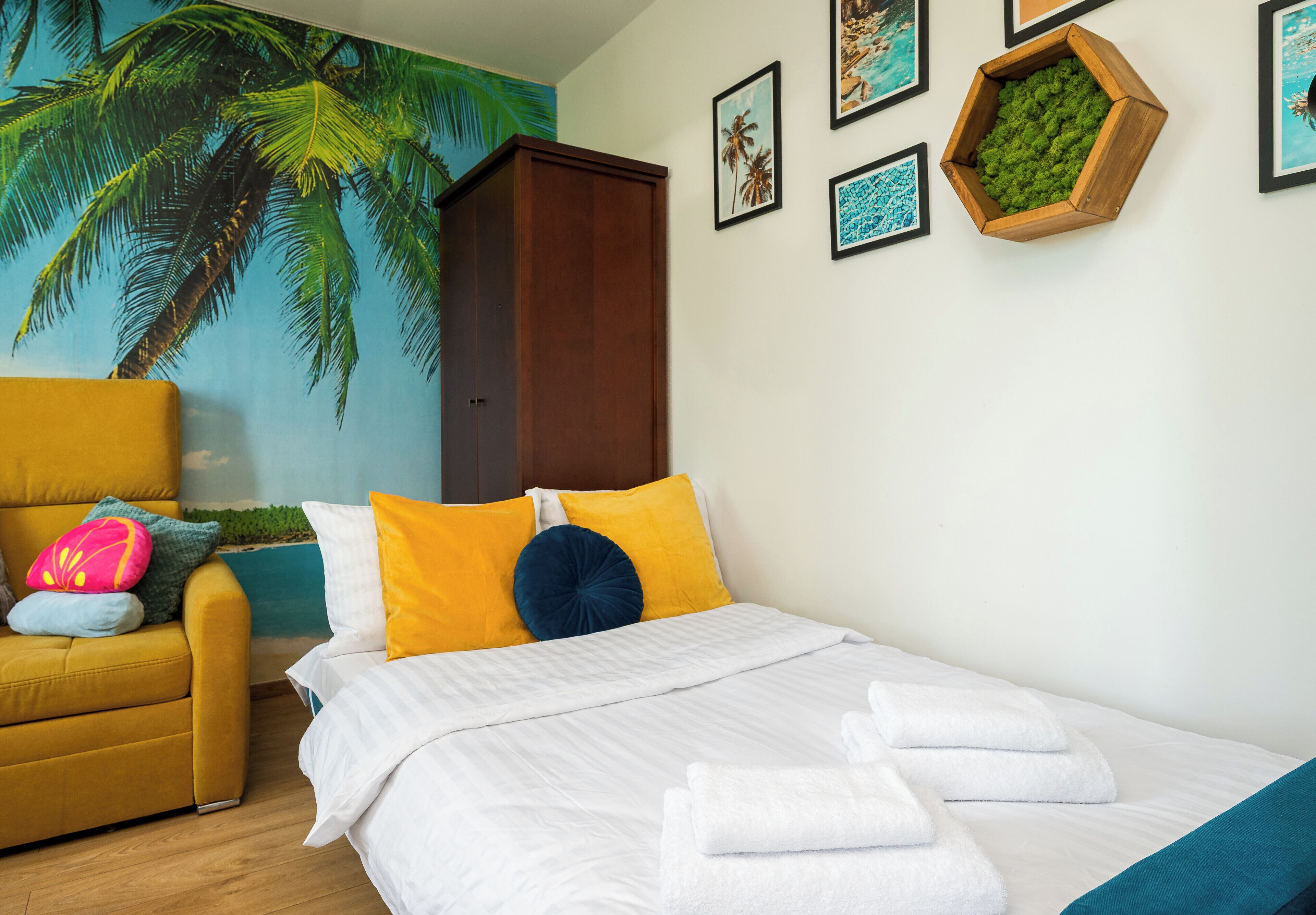 A bedroom with a bed, a yellow armchair, a mural of palm trees, and framed pictures on the wall.