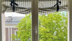 A window with a striped curtain tied back, revealing a view of a tree and a brick building. - Nowy Świat Sunny Side Apartment (Warszawa)