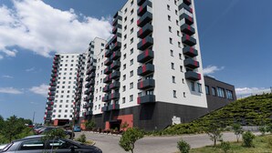 A modern high-rise building with multiple balconies, a flat roof, and a parking area in front.