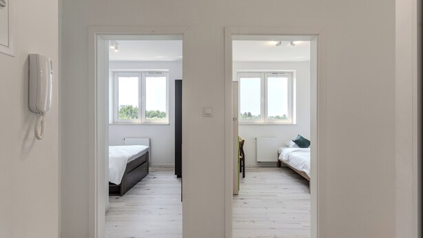 A hallway leading to two bedrooms with white walls, wooden flooring, and large windows.