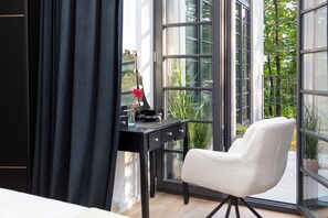 Ein Zimmer mit großem Fenster, einem Schreibtisch mit Blumenvase und einem Sessel.
