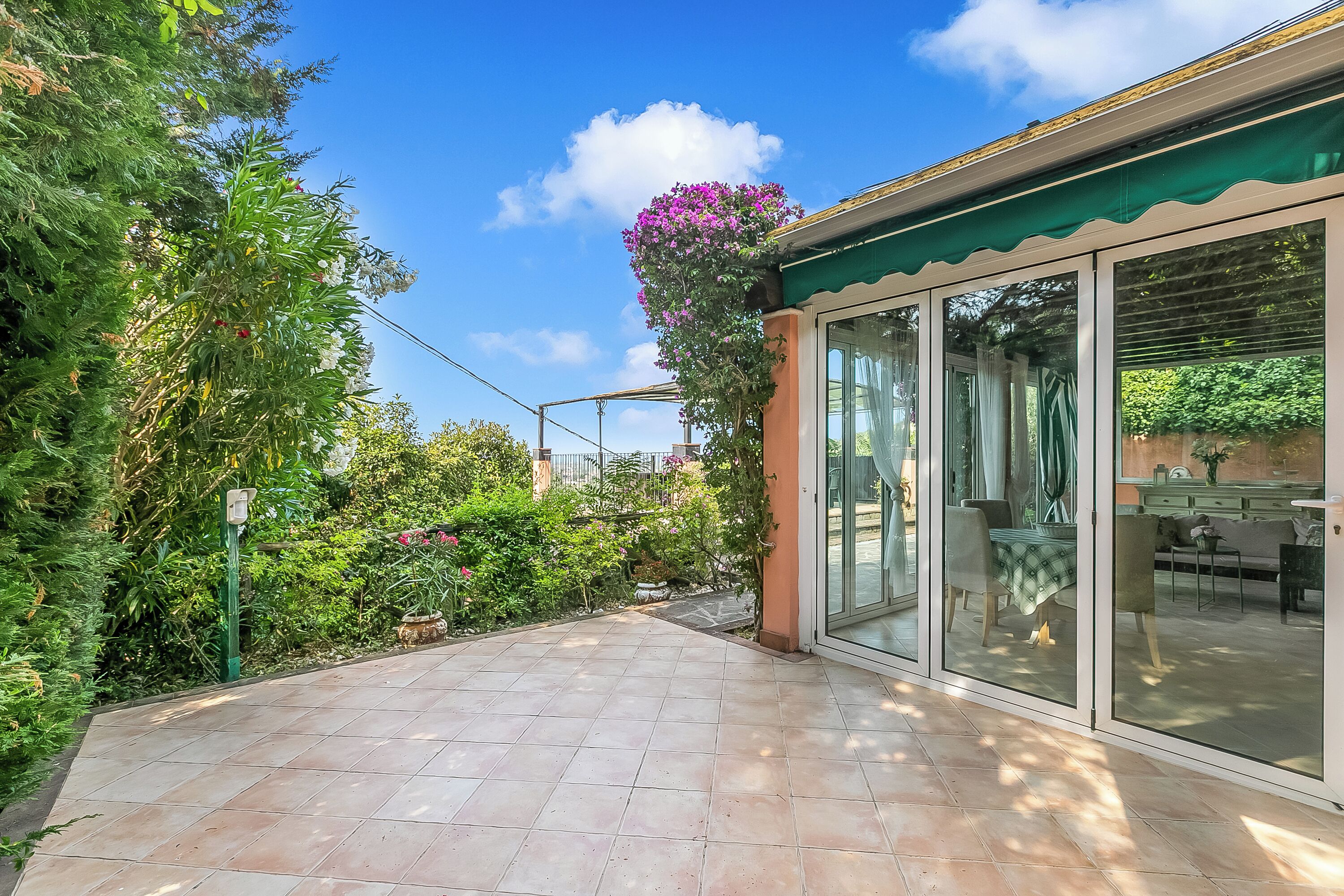 A patio with a tiled floor, a covered area with glass doors, and lush greenery.