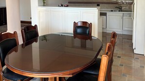 Une salle à manger avec une table en verre et des chaises en bois, un lustre, et une cuisine en arrière-plan.