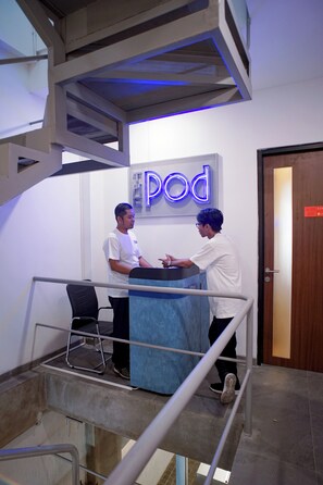 Zona de recepción con mostrador, un cartel que dice 'Pod' y una escalera con barandilla metálica.