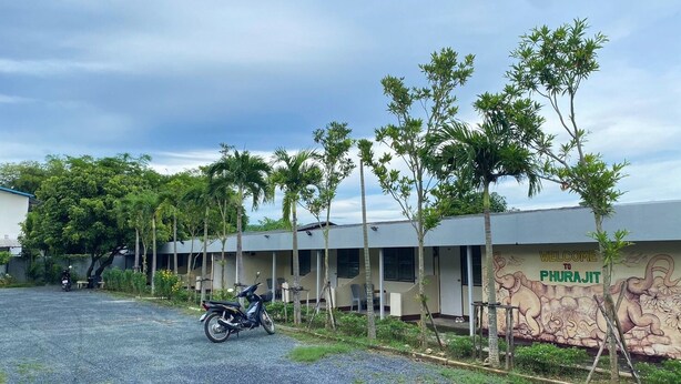Um edifício com um mural dando as boas-vindas aos visitantes em Phurajit, cercado por árvores e uma motocicleta estacionada.