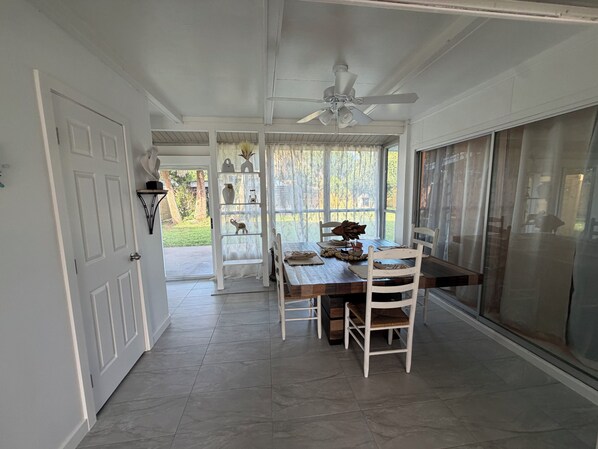 A dining area with a table and chairs, a ceiling fan, a window with a view of the outdoors, and a door leading to another room.