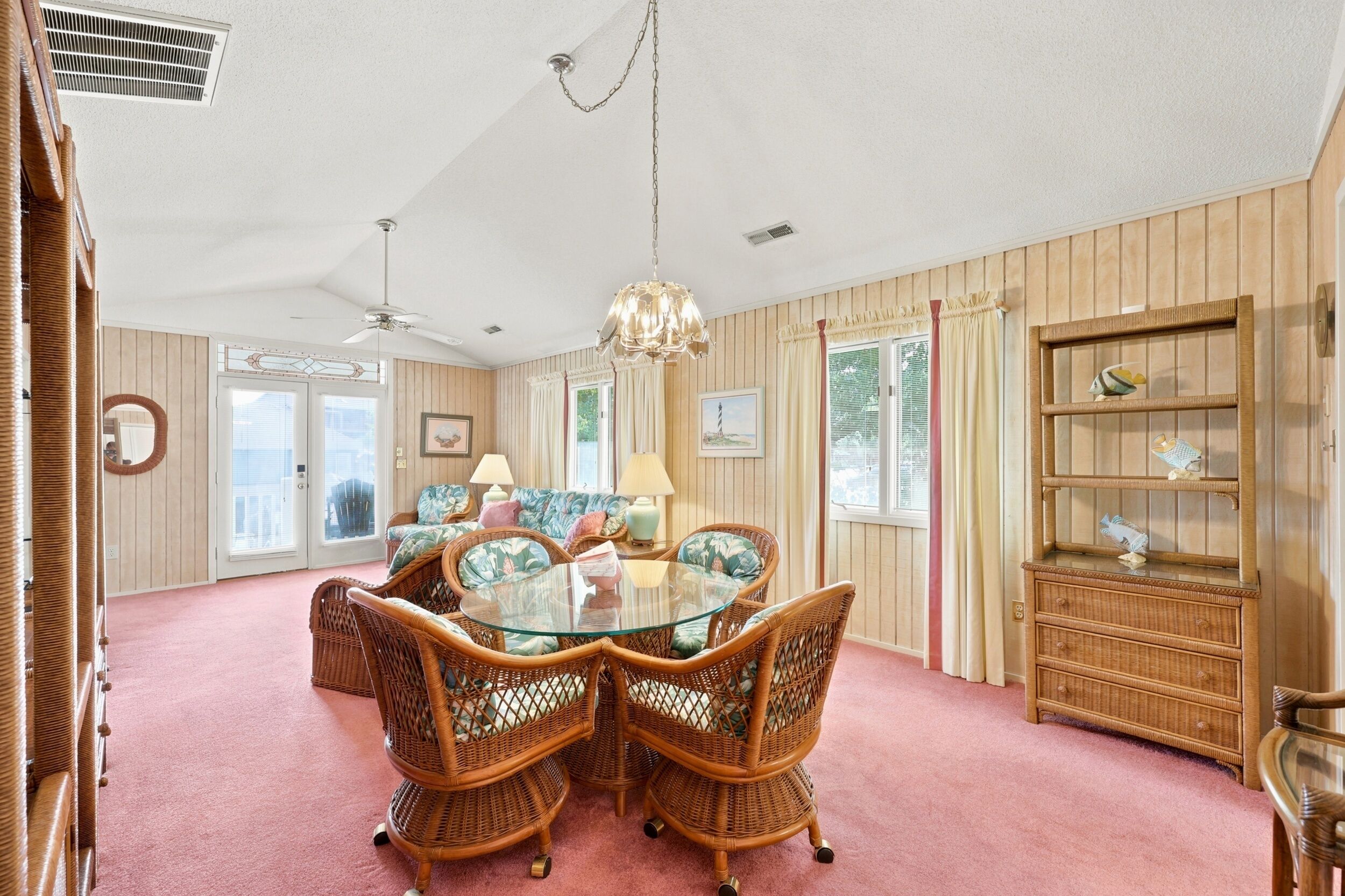 A room with a glass-top dining table surrounded by wicker chairs, a chandelier, and a wooden bookcase.