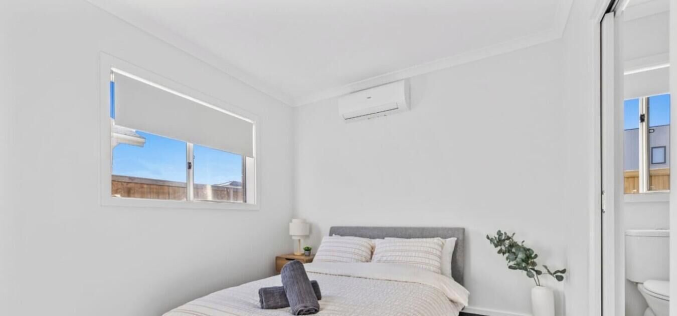 A bedroom with a bed, a bedside table with a lamp, a window, and a wall-mounted air conditioning unit.