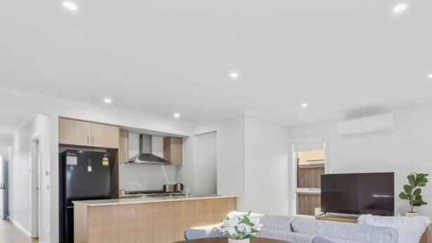 A modern living room with a sofa, a flat-screen TV, and a kitchen area with wooden cabinets and a black refrigerator. - Spacious Modern Retreat in Officer | Perfect for Families (Officer)
