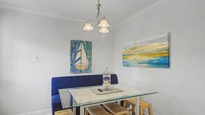 Zona pranzo con tavolo in vetro, sedie di legno, divano blu, lampadario e due quadri alle pareti.