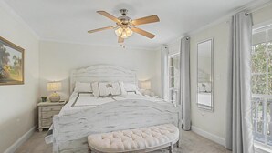 Una camera da letto con un letto, una ventola a soffitto, un comodino, una lampada, una finestra con tende e uno specchio.