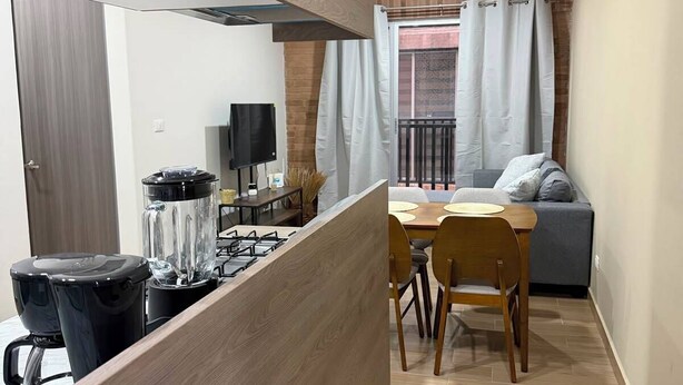 Cucina compatta con piano cottura a gas, frullatore e macchina per il caffè. È presente un'area pranzo con tavolo e sedie, e un divano grigio sullo sfondo.