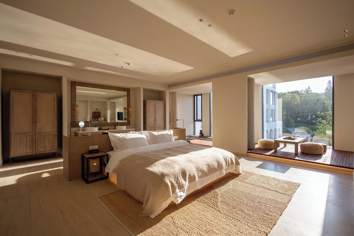 Une chambre à coucher moderne avec un grand lit, une table de chevet, un miroir et une vue sur l’extérieur.