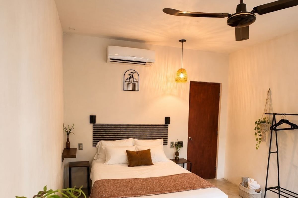 Un dormitorio con cama, mesitas de noche, ventilador de techo, aire acondicionado, lámpara colgante, puerta, planta y una pequeña mesa.