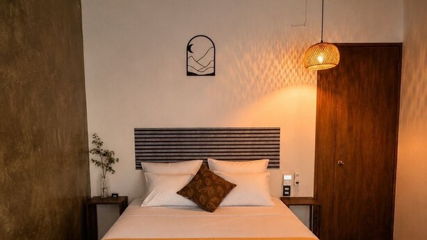 Una cama bien hecha con cabecera, una mesita de noche con lámpara y un reloj de pared.
