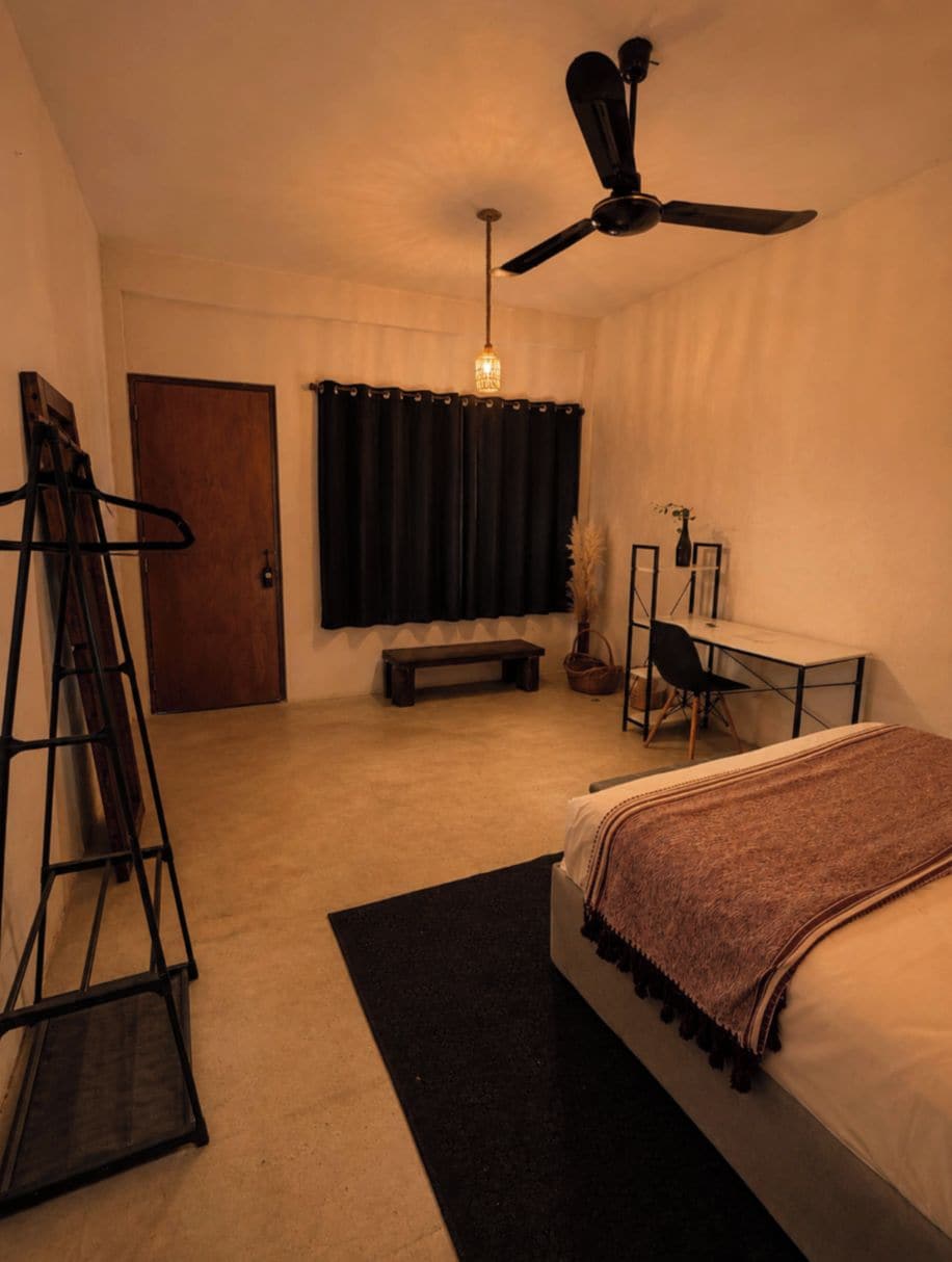 Un dormitorio con cama, escritorio, silla, ventilador de techo, puerta y ventana con cortinas.