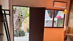Una habitación con piso de concreto, una cama con manta naranja, un escritorio con silla, una lámpara colgante y una puerta que da al exterior.