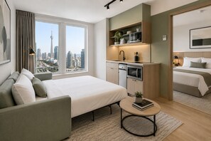 Ein modernes Hotelzimmer mit Bett, einer Sitzecke, einer kleinen Küchenzeile und Blick auf die Skyline der Stadt.