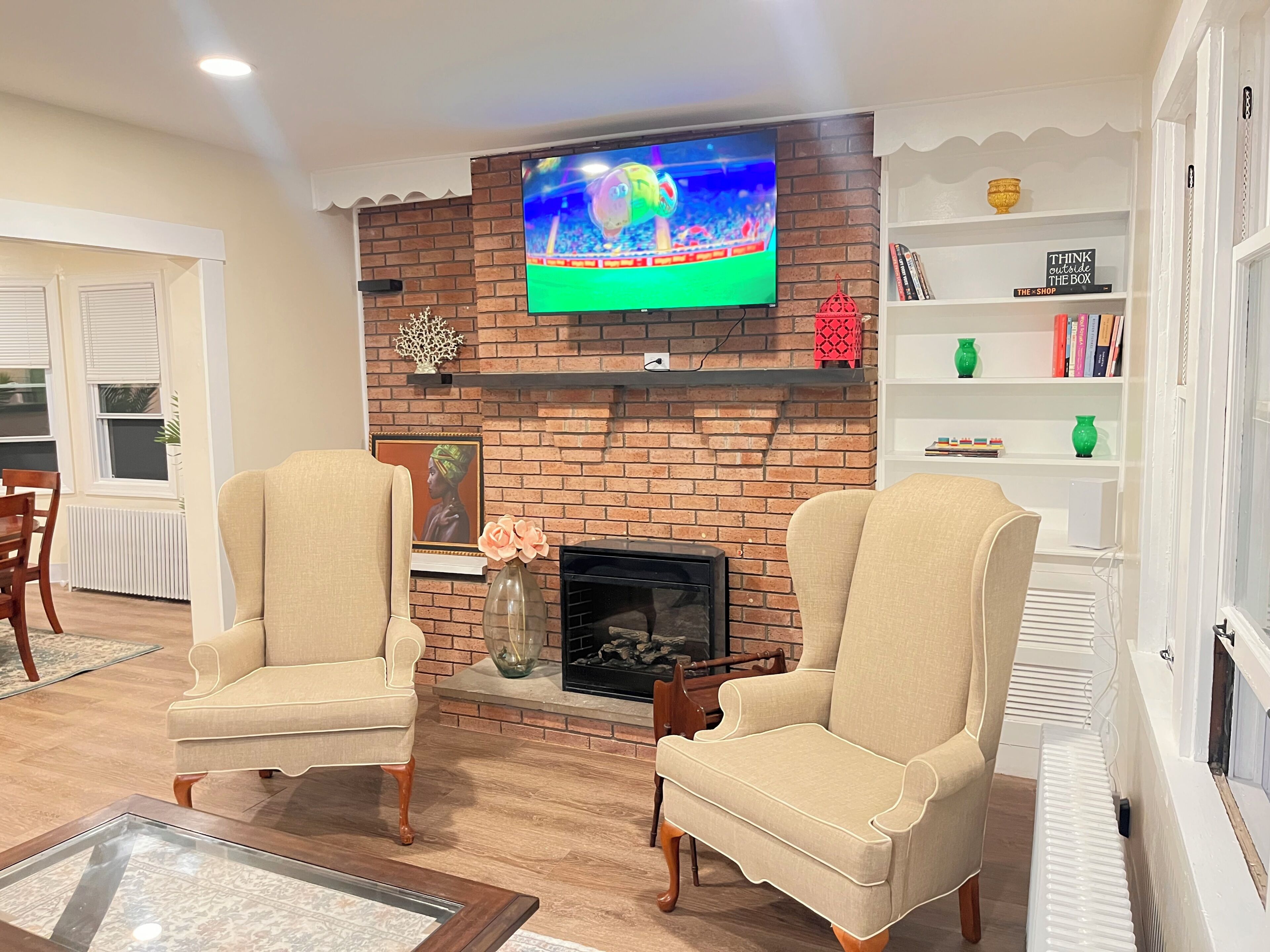 A living room with a brick fireplace, two armchairs, a television mounted on the wall, and a bookshelf.