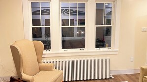 Ein Zimmer mit einem beige gefärbten Sessel, einem Heizkörper und einem großen Fenster mit vielen Sprossen.