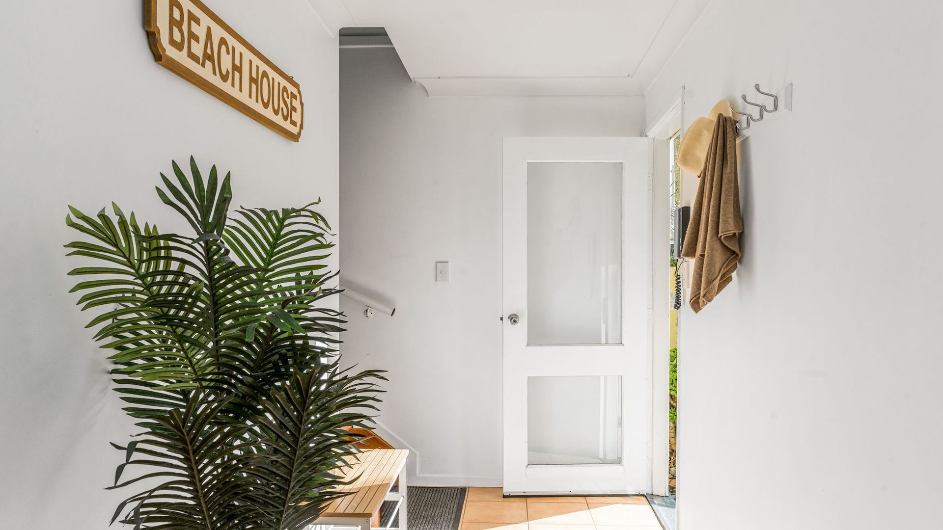 A hallway with a white door, a plant, a wooden sign reading 'BEACH HOUSE', and a towel rack.