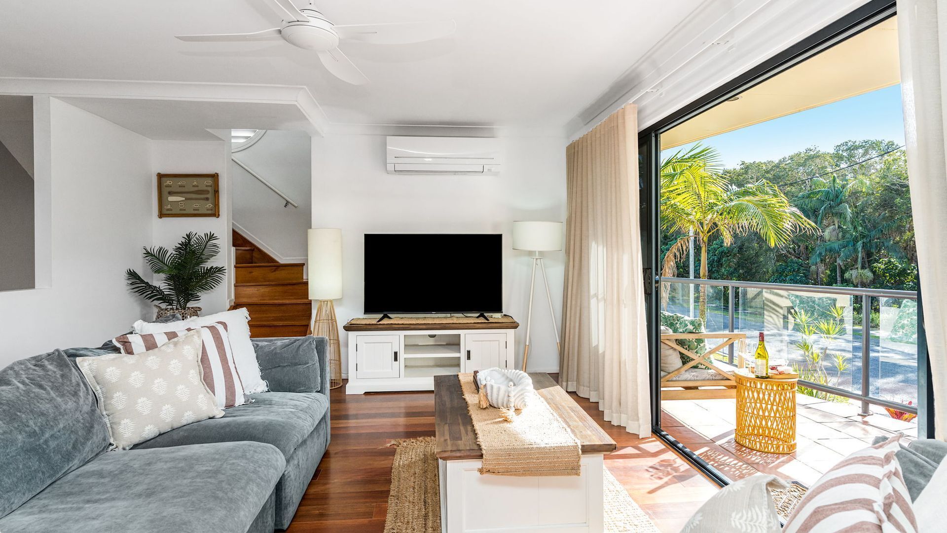 A modern living room with a flat-screen TV, grey sofa, wooden flooring, and a balcony with a view of greenery.