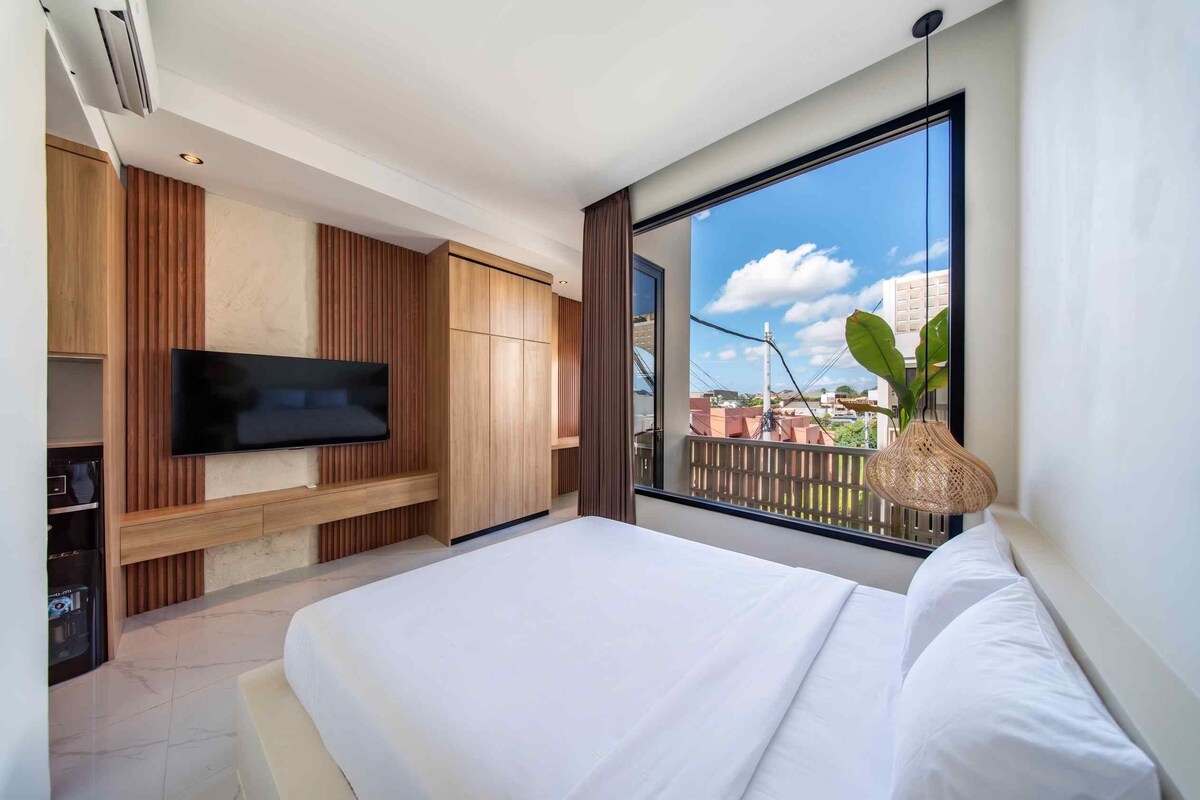 A modern bedroom with a large bed, a TV mounted on a wooden panel wall, and a balcony with a view of buildings.