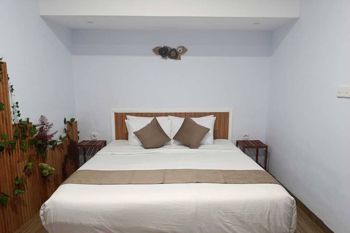 Un dormitorio con cama, cabecera de madera, paredes blancas, un adorno pequeño sobre la cama y una pared verde con plantas a la izquierda.