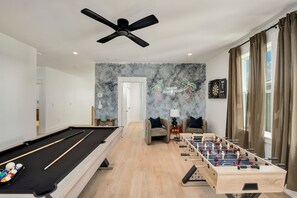 Ein Billardtisch, ein Tischfußballspiel und eine Dartscheibe in einem Raum mit Deckenventilator, Dartscheibe und einer Wand mit dem Schriftzug 'Das ist unser glücklicher Ort'.