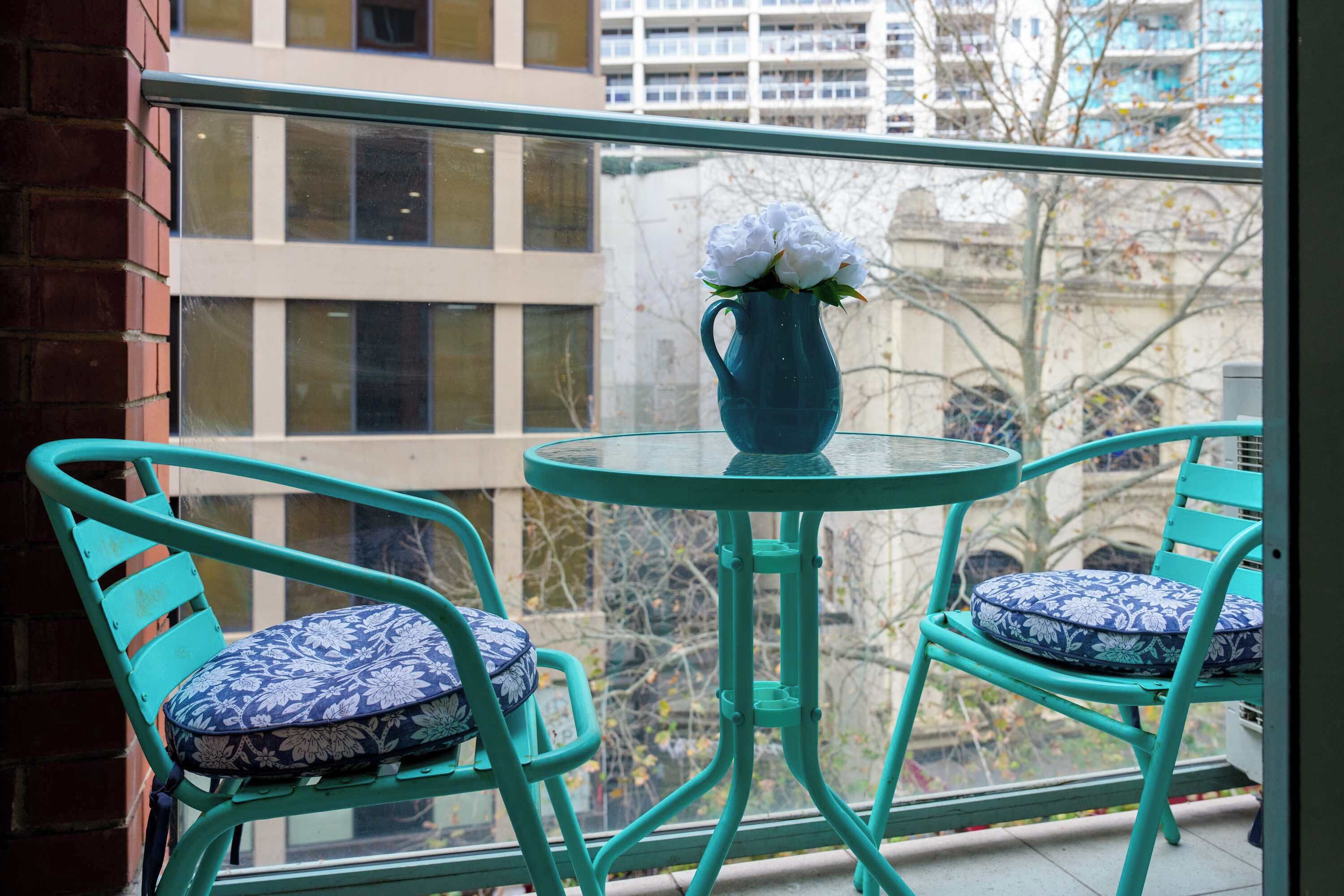 Step out onto the compact city balcony furnished with vibrant turquoise metal chairs with patterned blue seat cushions and a round glass bistro table holding a vase of white flowers.