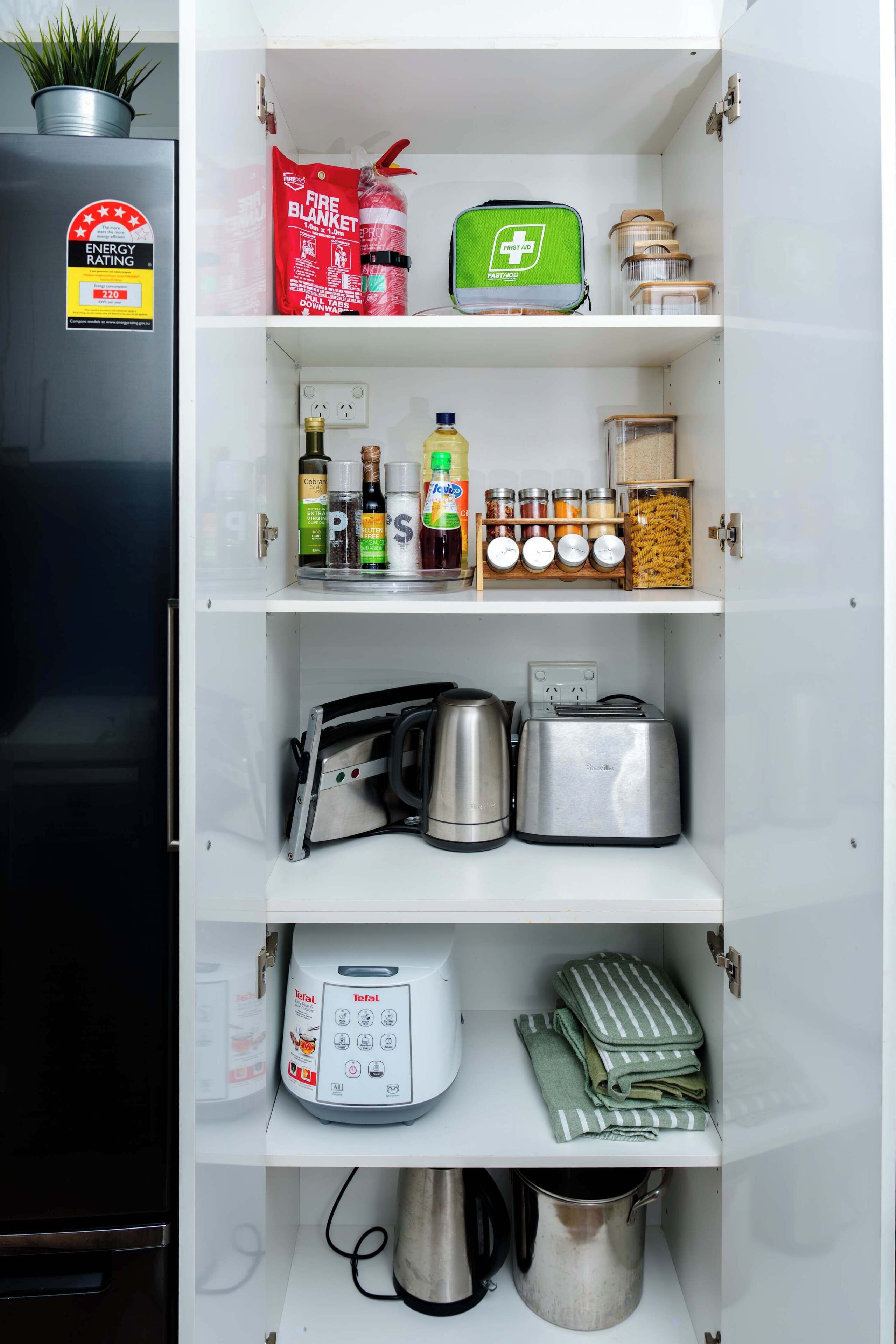 A kitchen with a refrigerator, a fire blanket, a first aid kit, cooking utensils, a toaster, a kettle, a food processor, and a coffee maker.