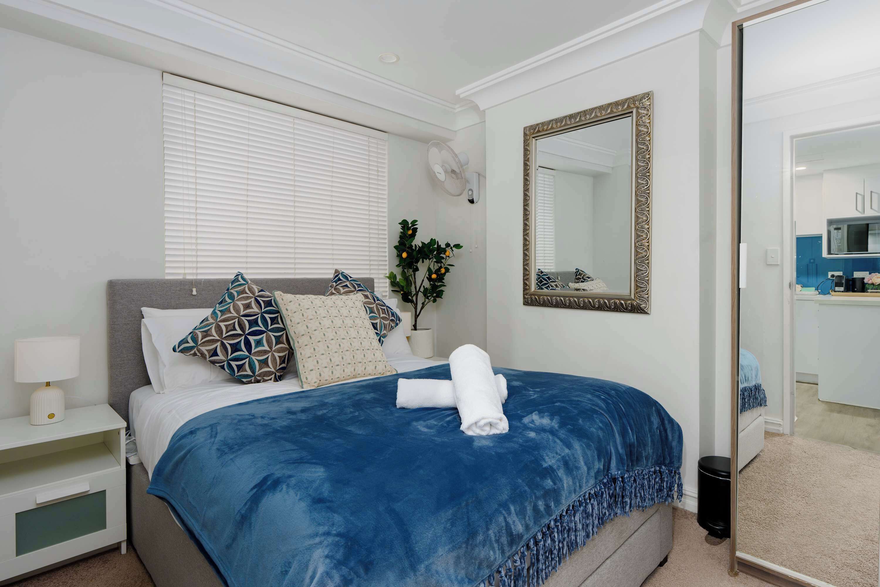 A hotel room with a bed, a bedside table with a lamp, a wall-mounted mirror, and a potted plant.