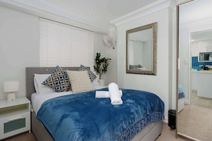 Une chambre d’hôtel avec un lit, une table de chevet avec une lampe, un miroir fixé au mur et une plante en pot.