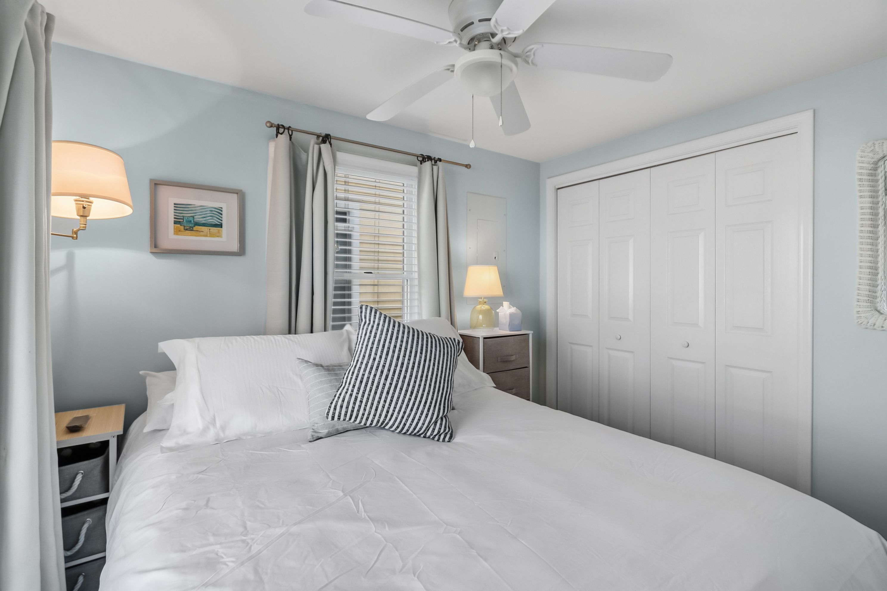 A bedroom with a bed, a ceiling fan, a window with blinds, a bedside lamp, and a door.