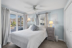 Ein Schlafzimmer mit Bett, Nachttisch, Lampe, Deckenventilator und Blick auf Gebäude durch das Fenster.
