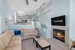 Una sala moderna con un sofá seccional color beige, un televisor de pantalla plana sobre la chimenea, y una cocina al fondo.